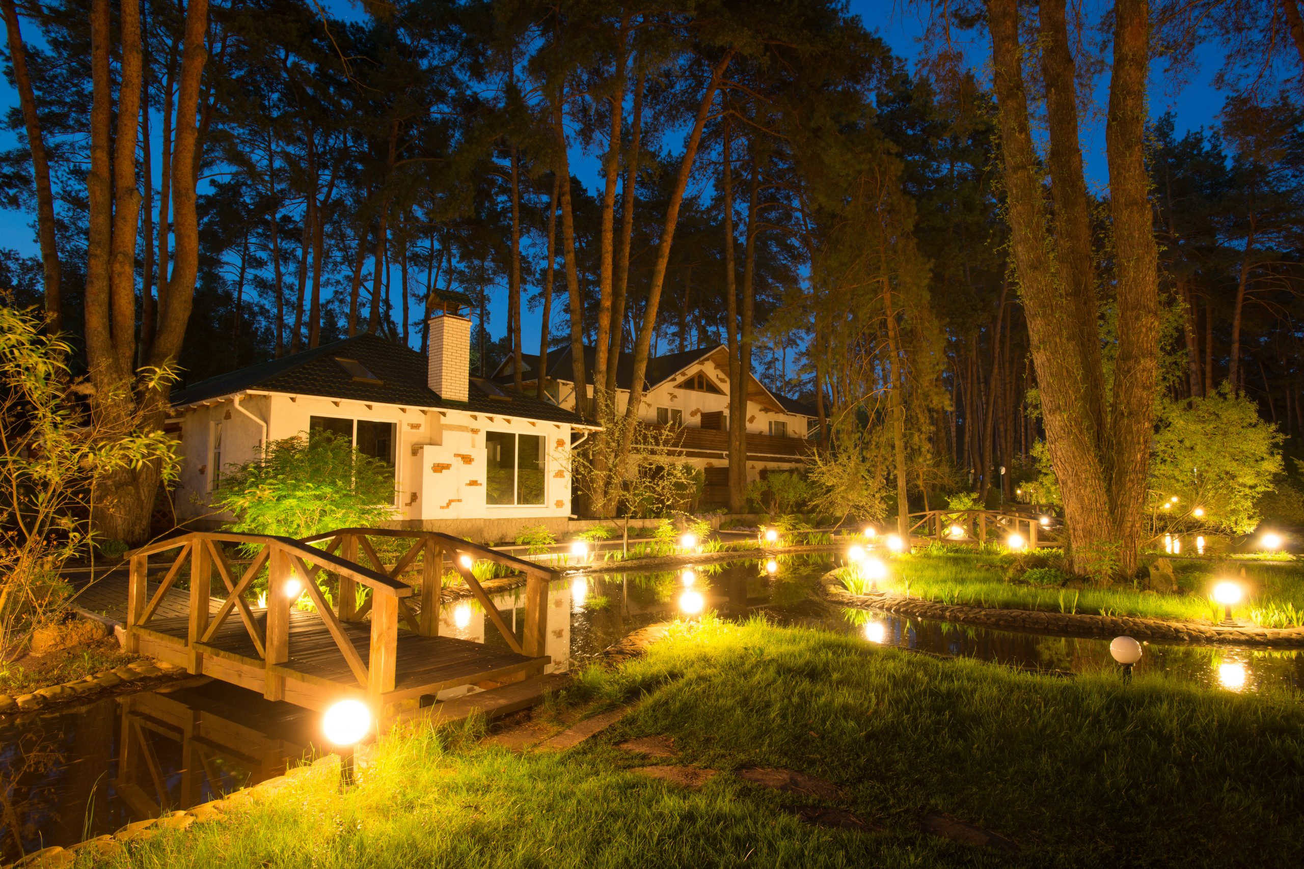A tranquil night scene with a well-lit garden by a pond and a cozy house amidst tall trees.